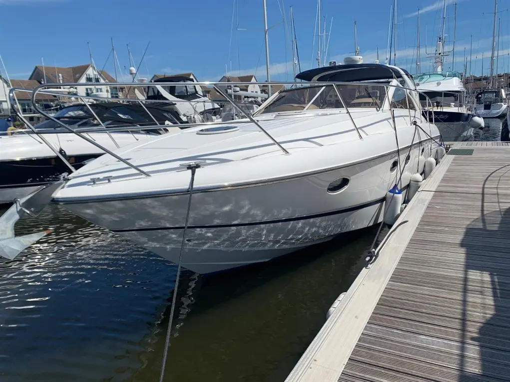A white boat at a marina, docked beside other vessels, with a clear blue sky and sunlight reflecting on the water.